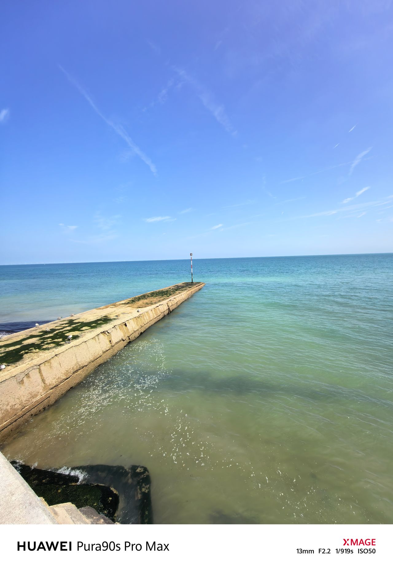 Ultra-wide view of a concrete groyne extending into calm turquoise water beneath a clear blue sky.