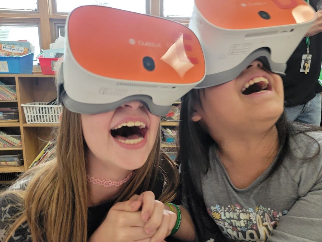 Two students with virtual reality headsets on react with joy to what they are seeing in the VR experience.