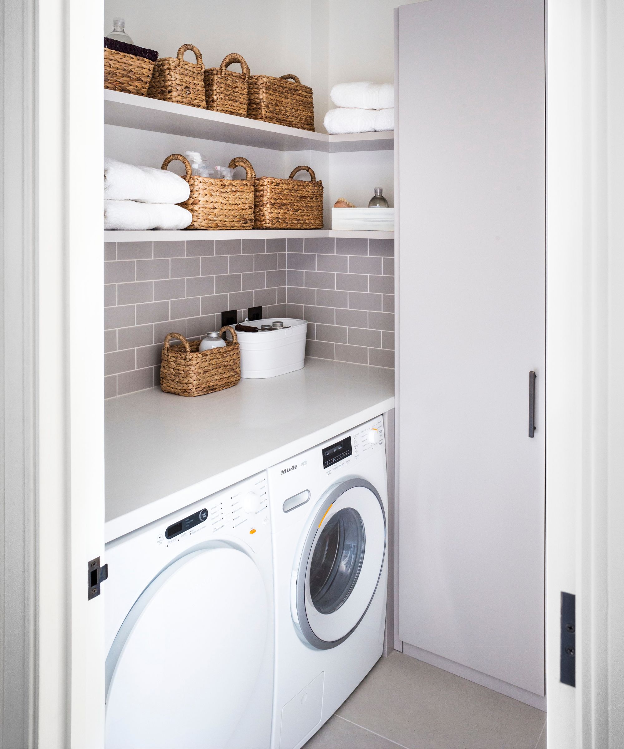 A clean and tidy purple tiled laundry room with a washing machine, tumble dryer, and shelves filled with wicker storage baskets and towels.