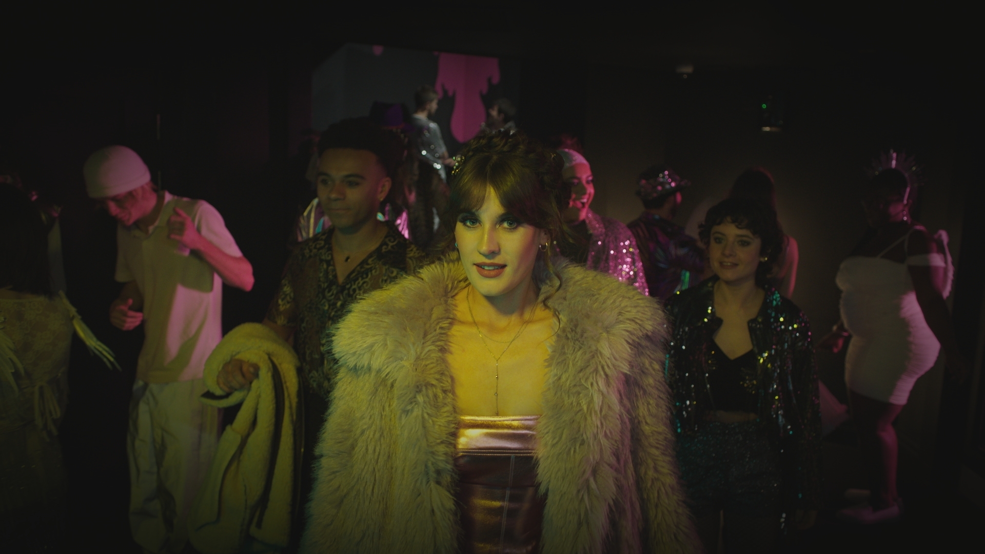 Jodie dressed in a glamorous shiny top with a big furry coat on looking at the camera while clubbers dance around her. Indie and Teddy can be seen smiling excitedly behind her.