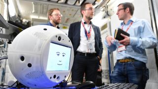 A close-up of CIMON, the Crew Interactive Mobile Companion, an A.I.-equipped robot that is the first of its kind to fly in space. In the background (from left to right) are: CIMON project manager Christian Karrasch; Till Eisenberg CIMON project lead at Airbus; and Christoph Kossl, Airbus software systems engineer.