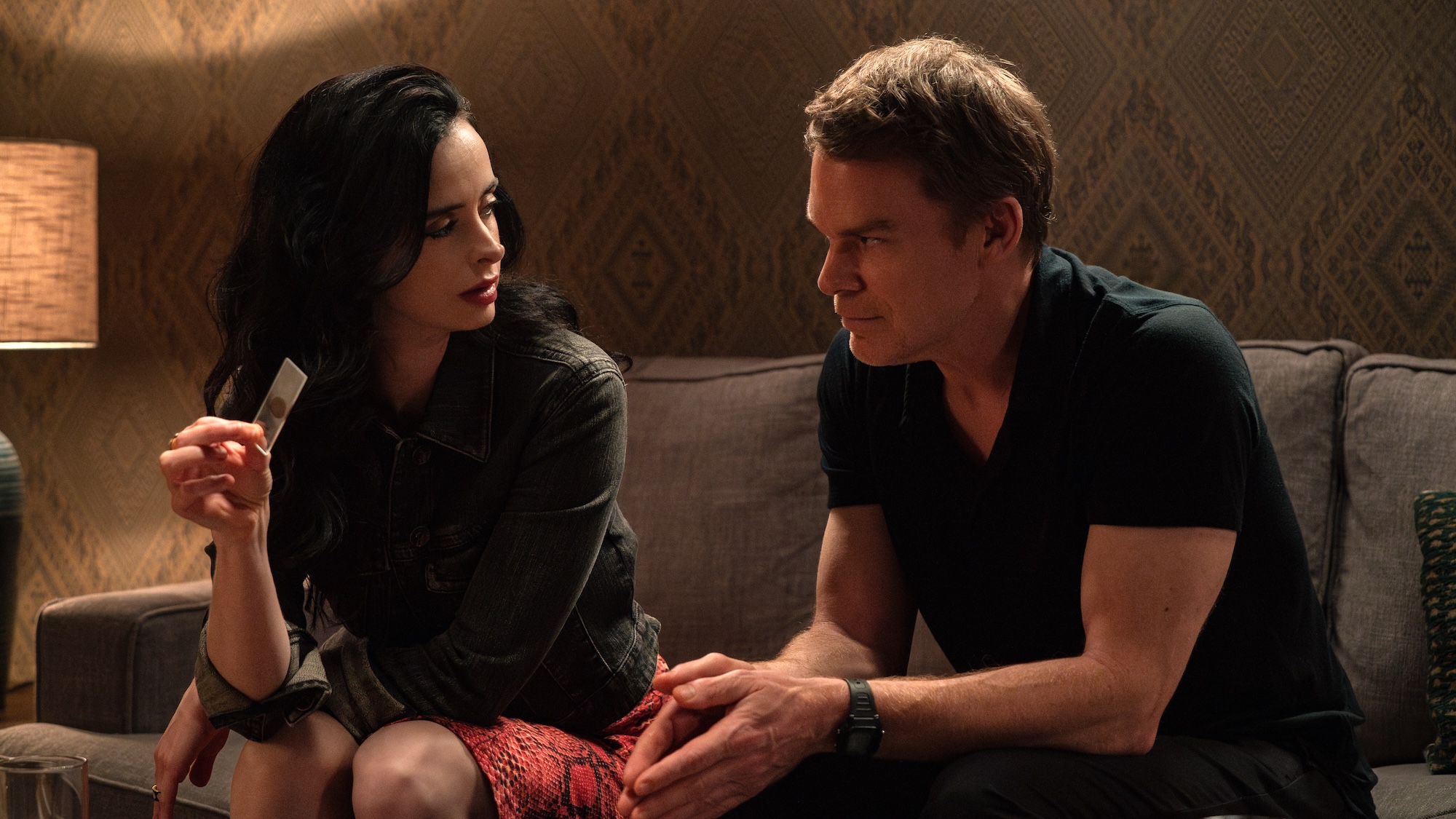 Michael C. Hall's Dexter in short sleeve shirt sitting on a couch next to hallucination of Krysten Ritter's Mia holding one of his blood slides in Dexter: Resurrection Season 2