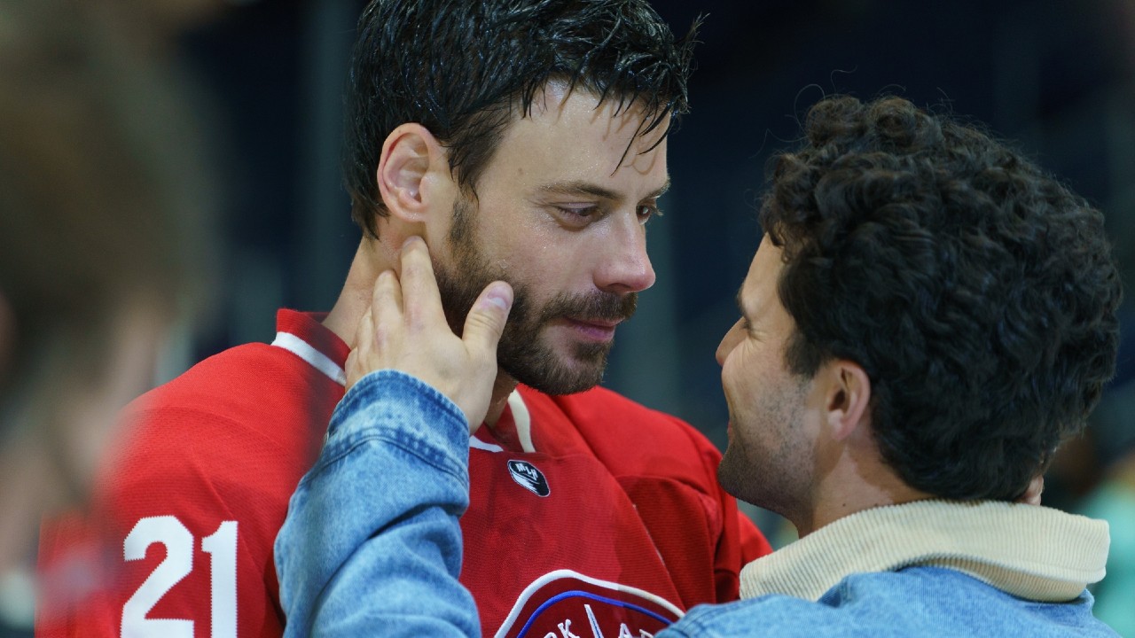 Scott, in his hockey gear, looking down at Kip as Kip touches his face.