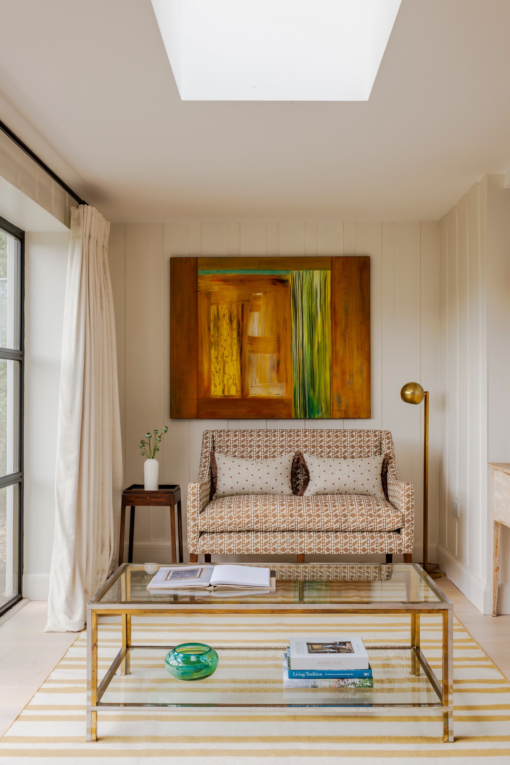 a small patterned sofa next to a side table and floor lamp with a glass coffee table on top of a striped rug in a small room with beige curtains and a skylight