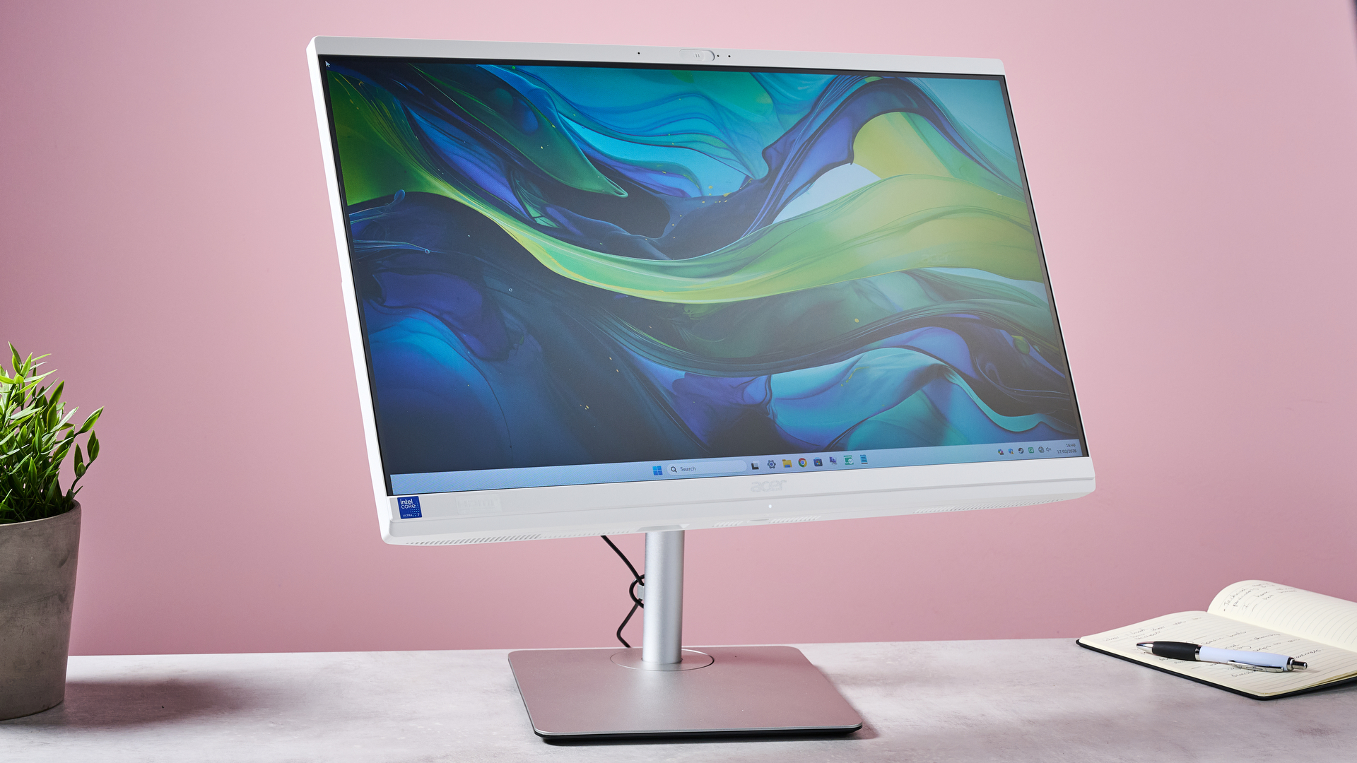 Three-quarter view of Acer Aspire C24 with display tilted upwards, on desk with plant and note pad against a pink background