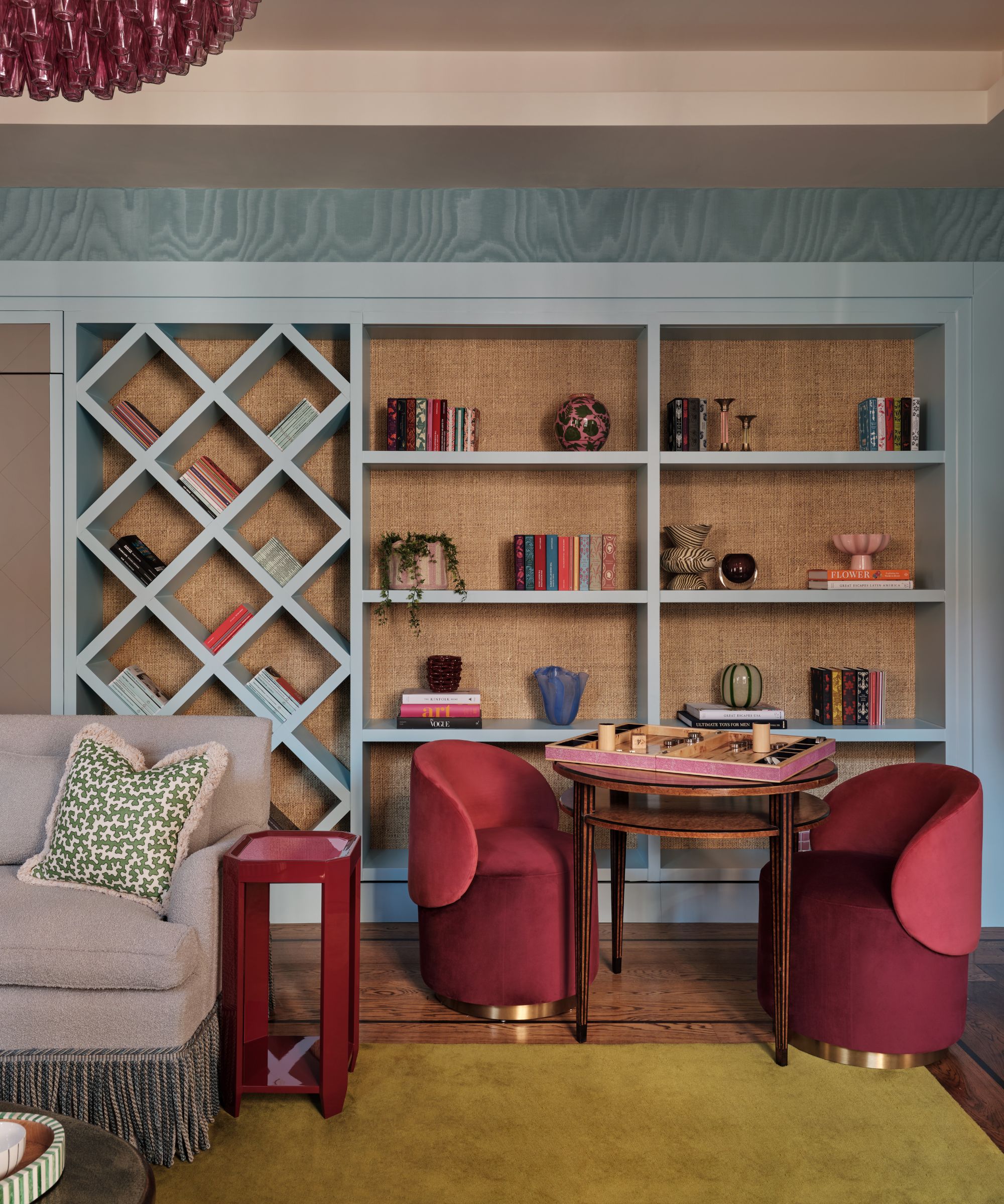 a blue moire wallpapered library with blue grasscloth covered bookcases, small pink velvet chairs with a small games table