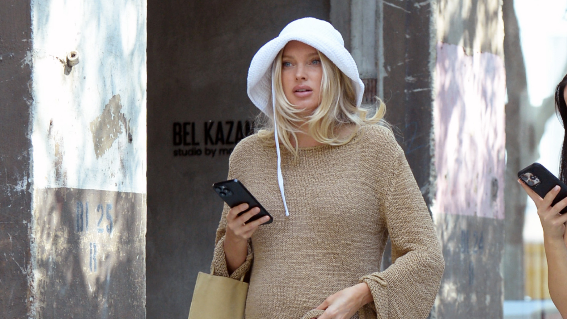 Elsa Hosk wearing a white bucket hat, tan summer sweater, white bermuda shorts, flip-flops, and a The Row Cecily Mini Tote Bag