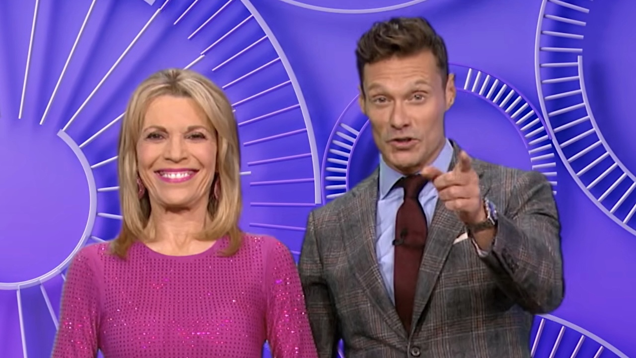 Vanna White smiling in fuchsa dress standing next to Ryan Seacrest pointing at camera on Wheel of Fortune