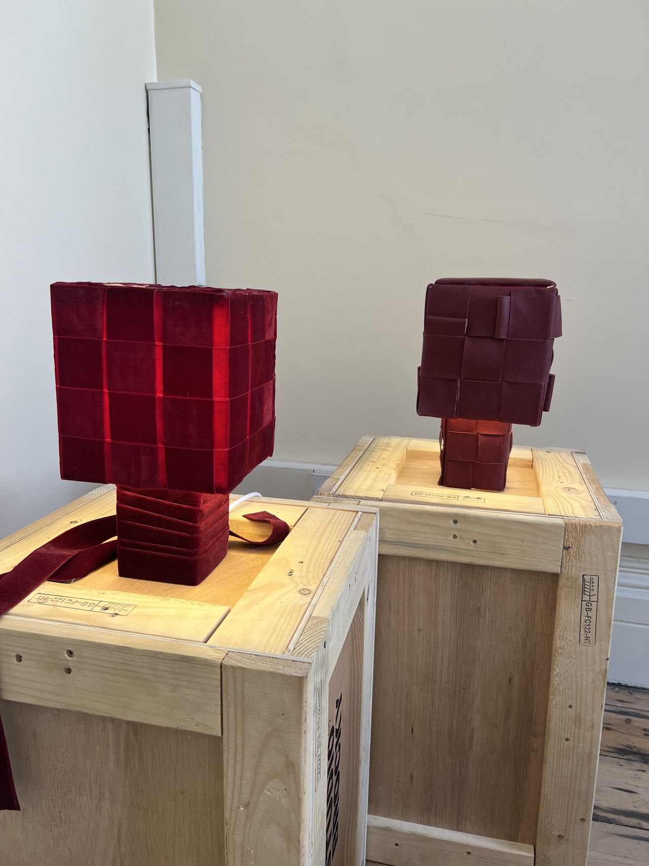 Image of two red lamps on wooden crates. One is woven from velvet and the other is woven from leather.