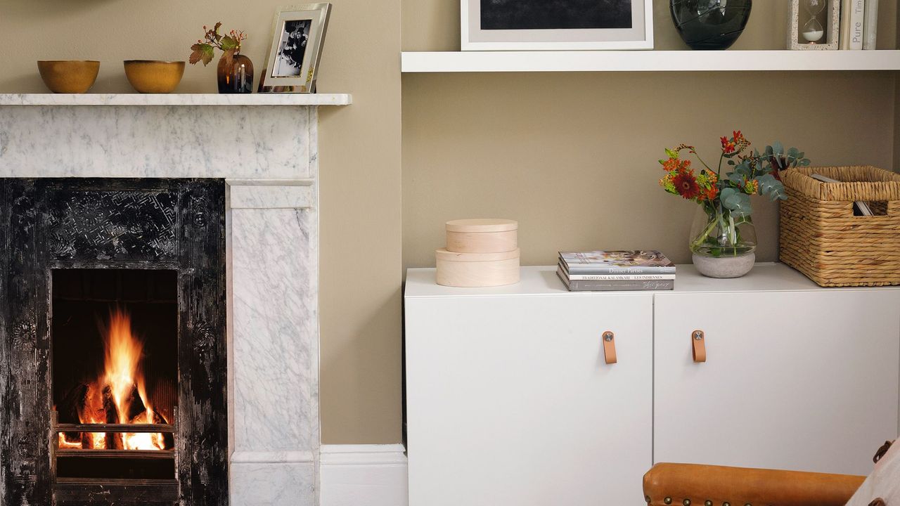 Living room with a lit fire in a traditional fireplace, shelves and a cupboard, a large leather armchair and cosy painted walls