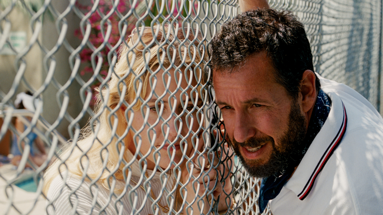 Greta Gerwig and Adam Sandler share a moment through a chain link fence in Jay Kelly.