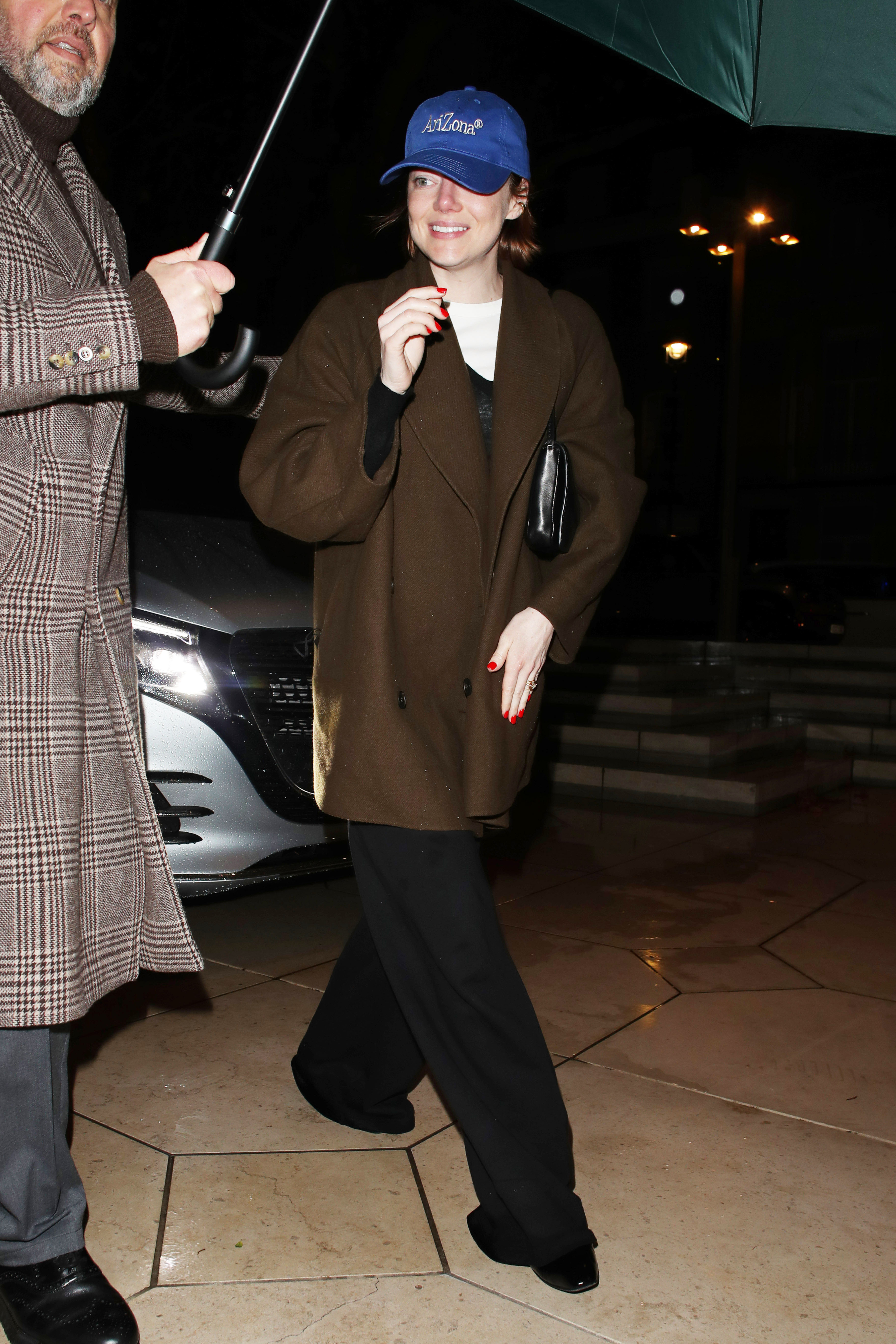 Emma Stone wearing a brown coat, black pants, and a blue baseball cap