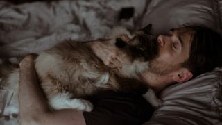 An owner is asleep in bed, hugging a Ragdoll cat that is peacefully sleeping on top of them.