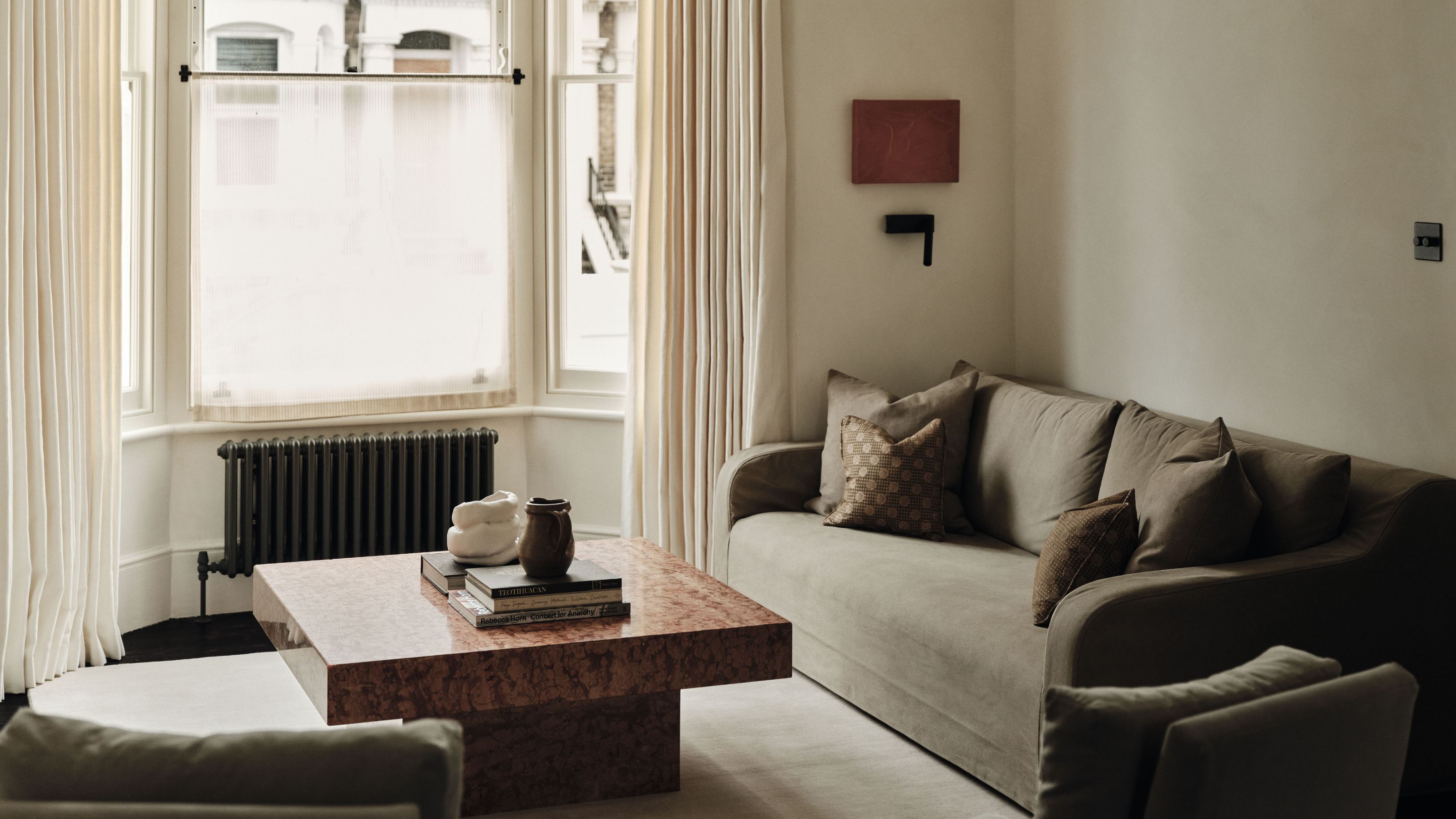 A neutral, minimalist living room by Studio Jey featuring a pink marble coffee table, a soft grey sofa, and a large bay window with cream drapery.