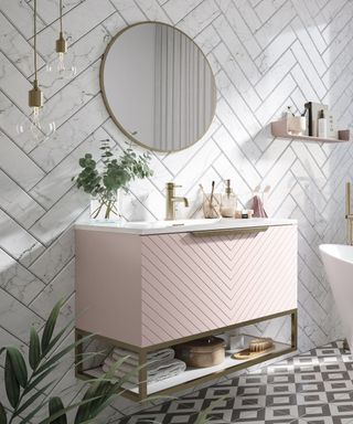 A bathroom with white marble herringbone tiles, a gold circular mirror and two bulbs, and a floating vanity with a light pink base and gold faucet