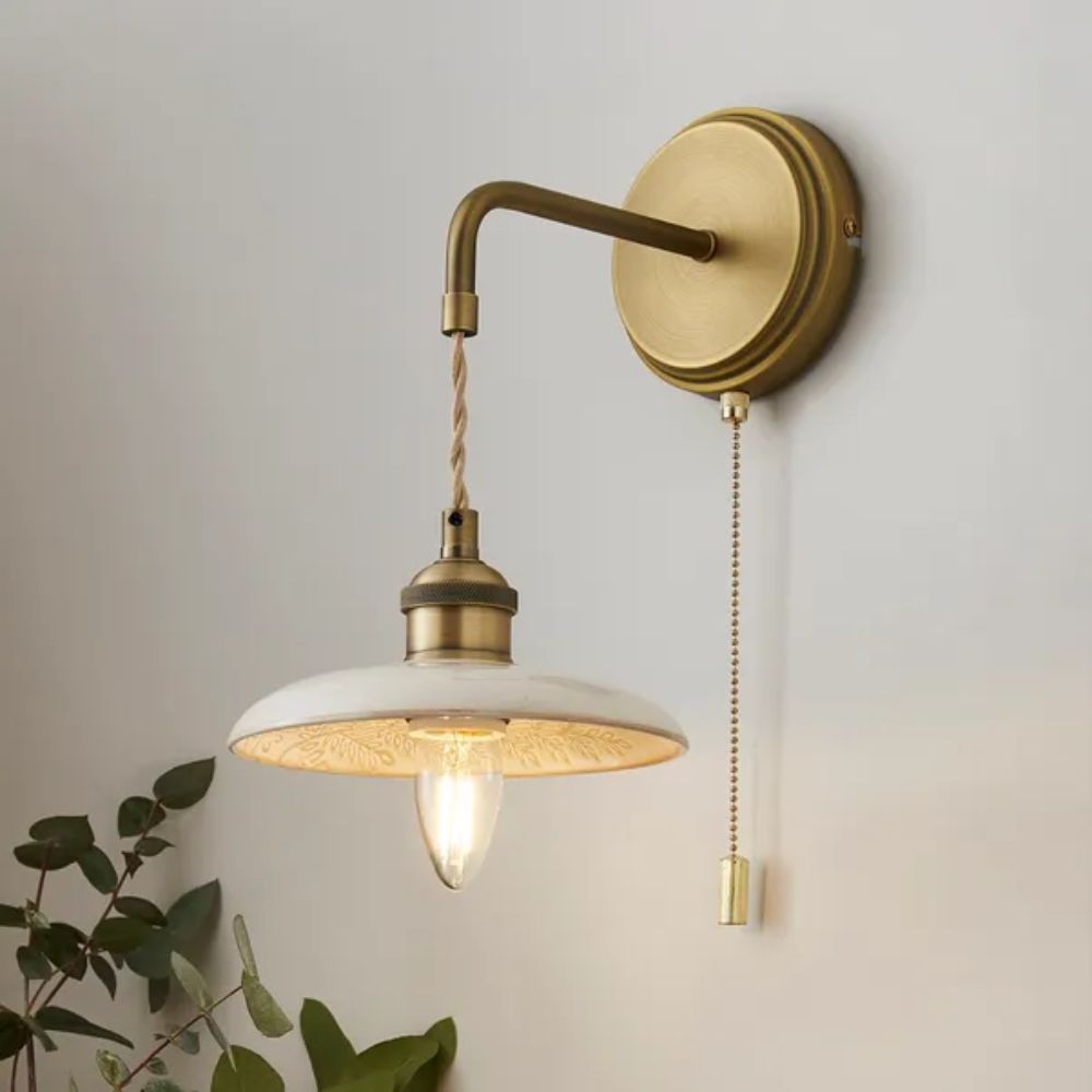 A warm brass wall light with a curved arm and pull chain switch, featuring a shallow white metal shade and exposed bulb.