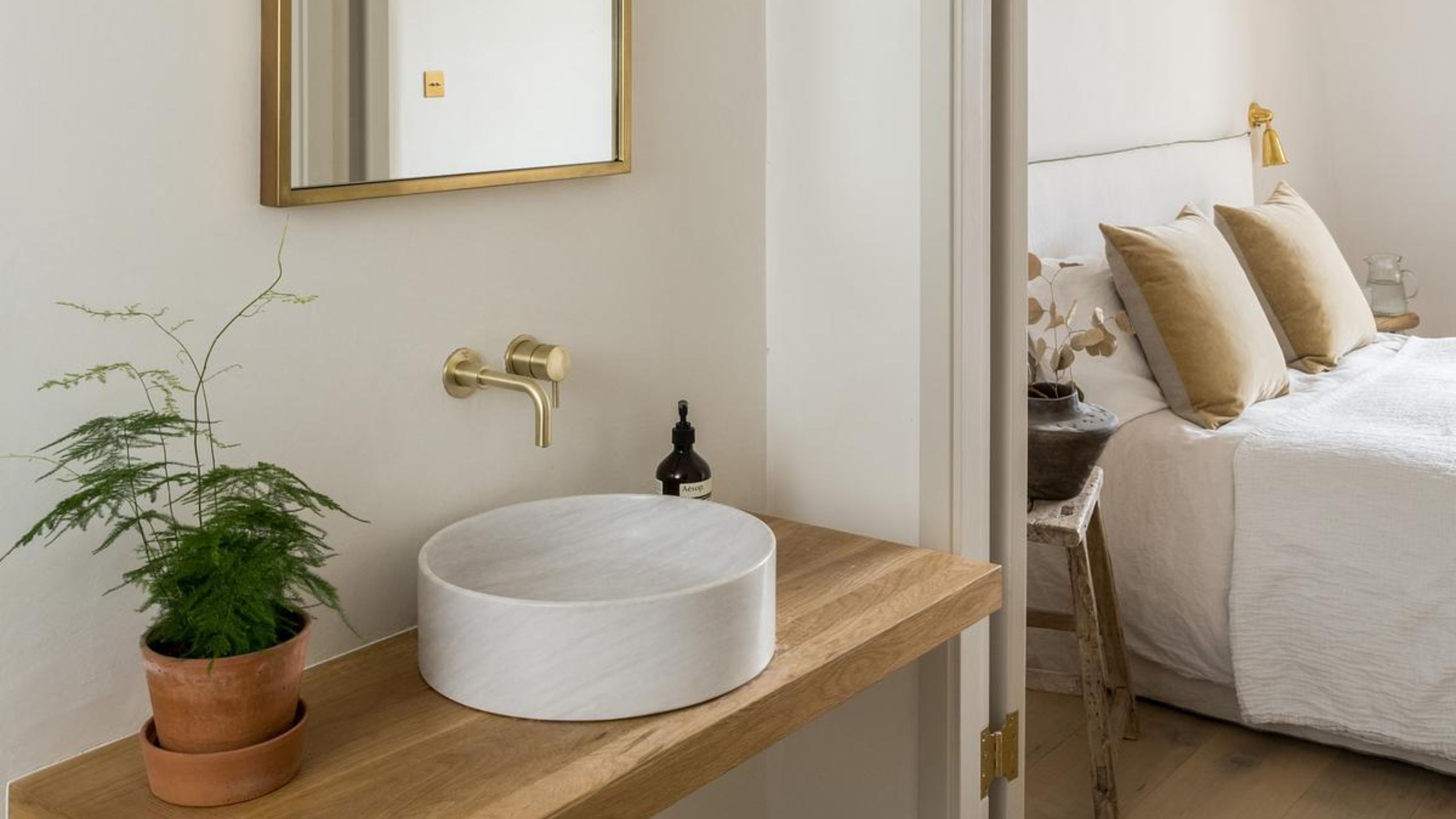 An ensuite with the view of a bedroom that has cushions on top as well as a view of the bathroom sink, countertop and mirror. On the countertop is handwash and a potted plant. 