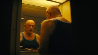 Close up of an older man wearing a dark vest whilst looking in a dimly lit bathroom mirror at his reflection.