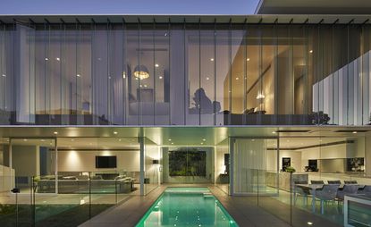 The exterior of a residential glass house on 2 floors with a pool. With the curtains open, the interior- lounge, kitchen and dining area are visible on the bottom floor and the bedroom visible on the top floor