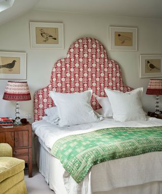 Cream painted bedroom with large, red patterned headboard, white linen with green patterned through, four framed images above bed, matching patterned bedside table lamps