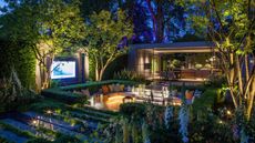 A modern garden at dusk with a conversation pit with lighting integrated into the seating, up-lit trees, and a view into a garden room with large windows