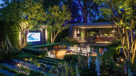 A modern garden at dusk with a conversation pit with lighting integrated into the seating, up-lit trees, and a view into a garden room with large windows