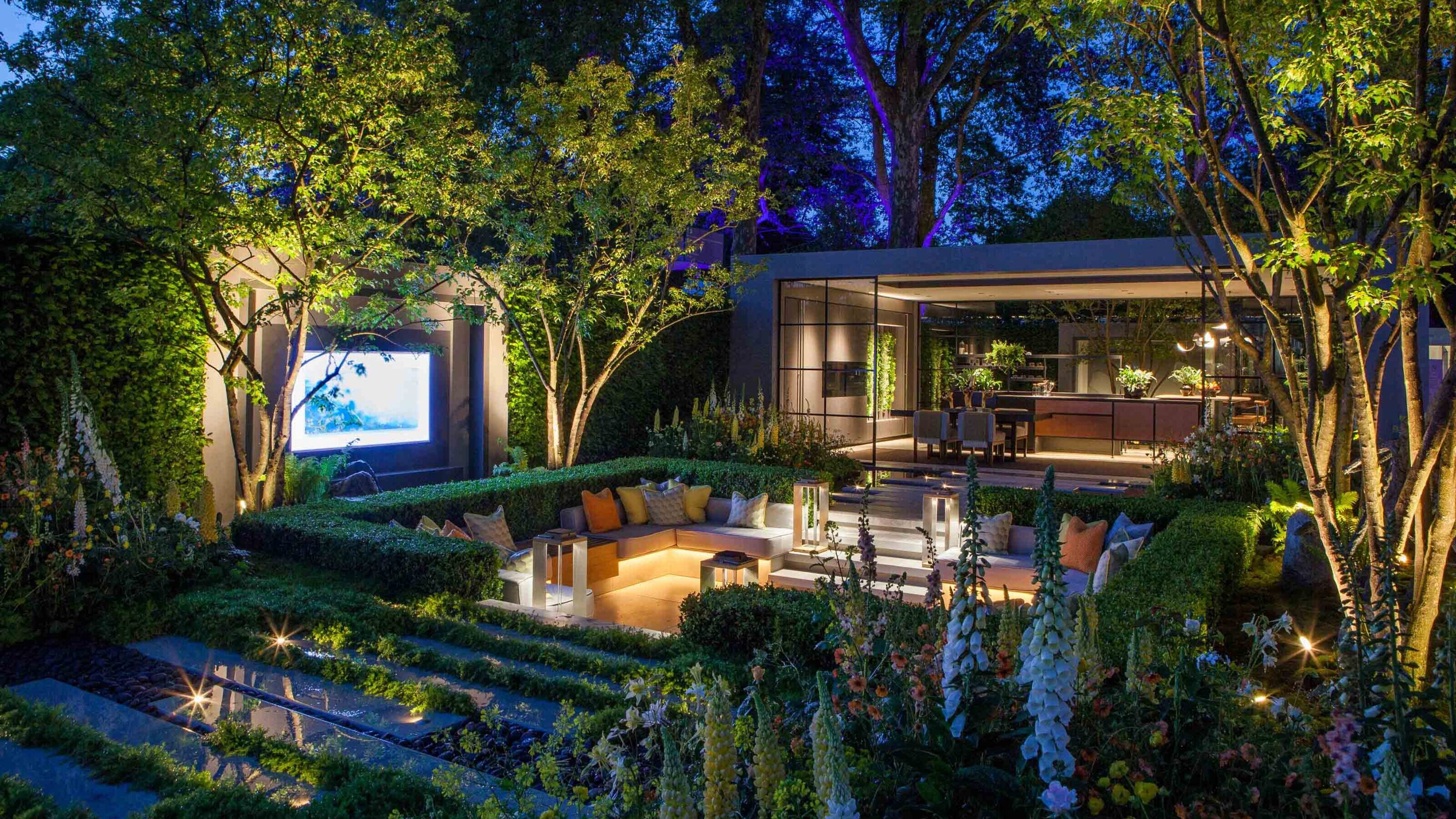 A modern garden at dusk with a conversation pit with lighting integrated into the seating, up-lit trees, and a view into a garden room with large windows