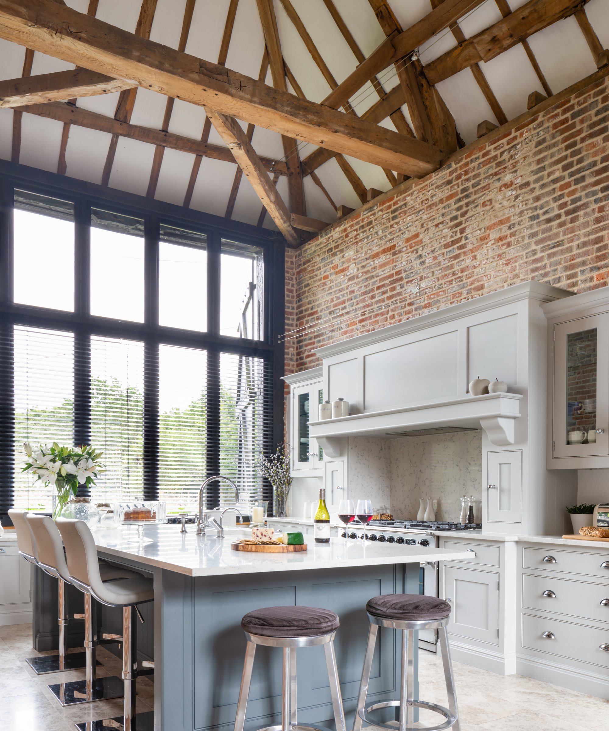 Open plan kitchen ideas in a tall beamed space with exposed brick wall, pale gray cabinetry and a kitchen island.
