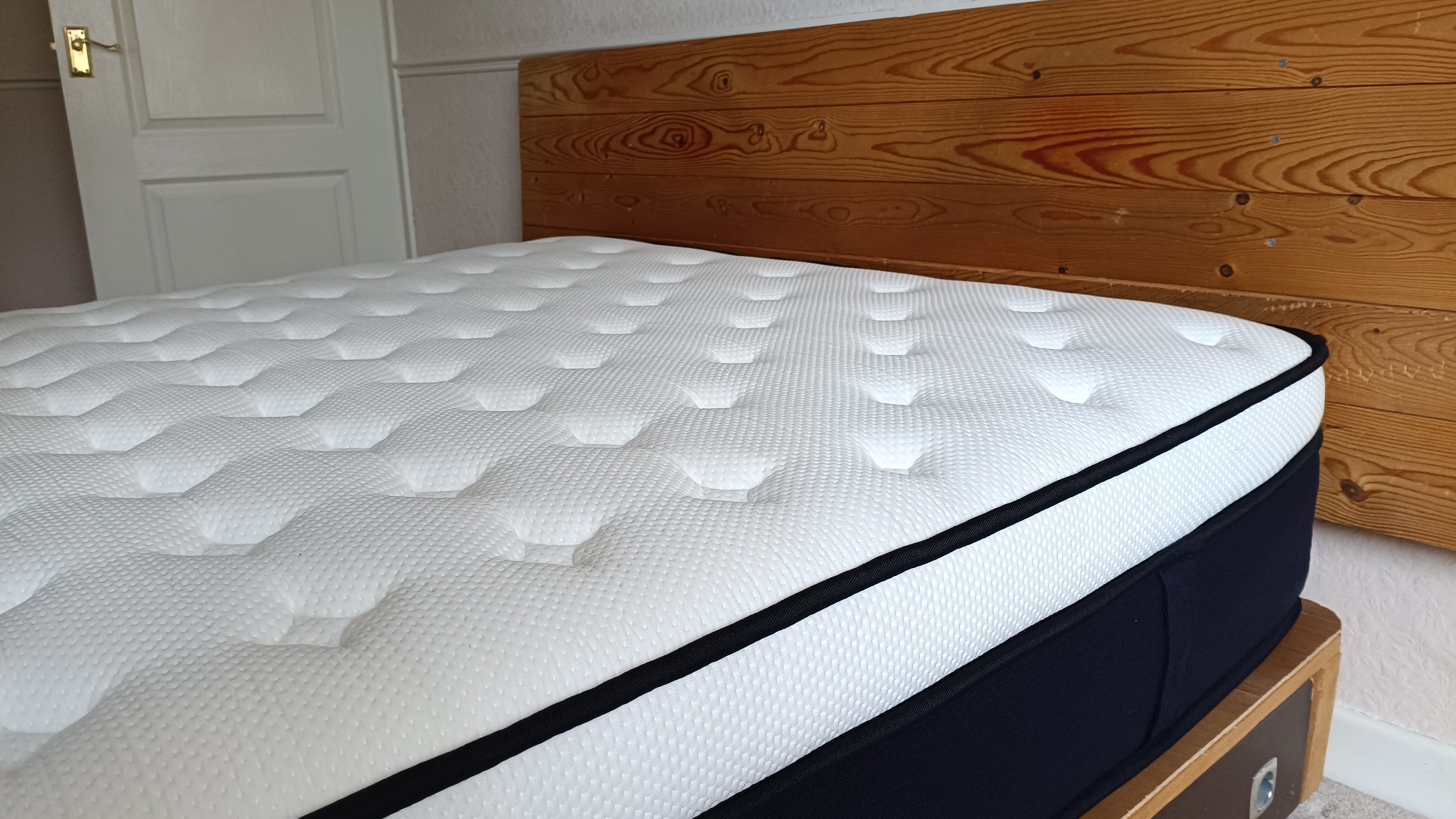 This wide-angle view highlights the mattress's thick profile and tufted white top surface set against a rustic wooden headboard.