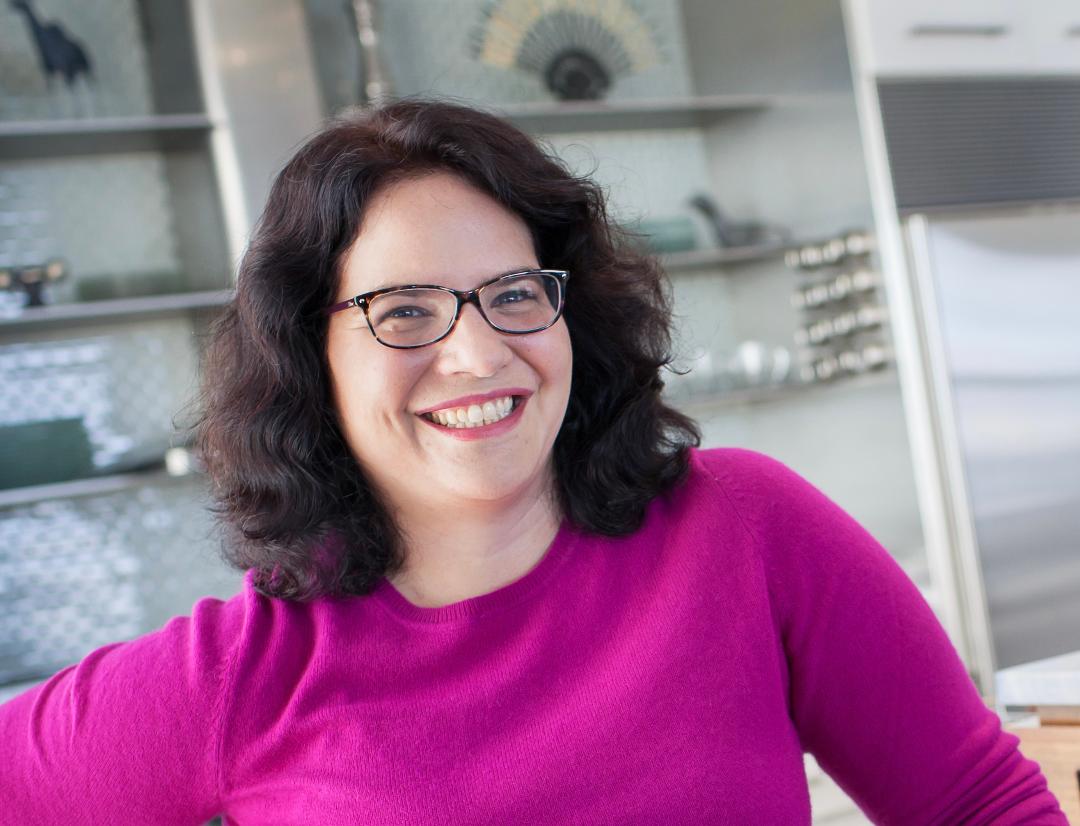 Amanda Wiss, Professional organizer and founder of Urban City, a home organizing company based in NYC. Also the founder of home staging studio, Urban Staging, a woman with dark hair wearing glasses and a bright pink jumper, against blurred shelves.