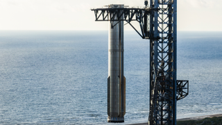 closeup of a silver rocket booster held above ground by the mechanical arms of its launch tower, with the ocean in the background