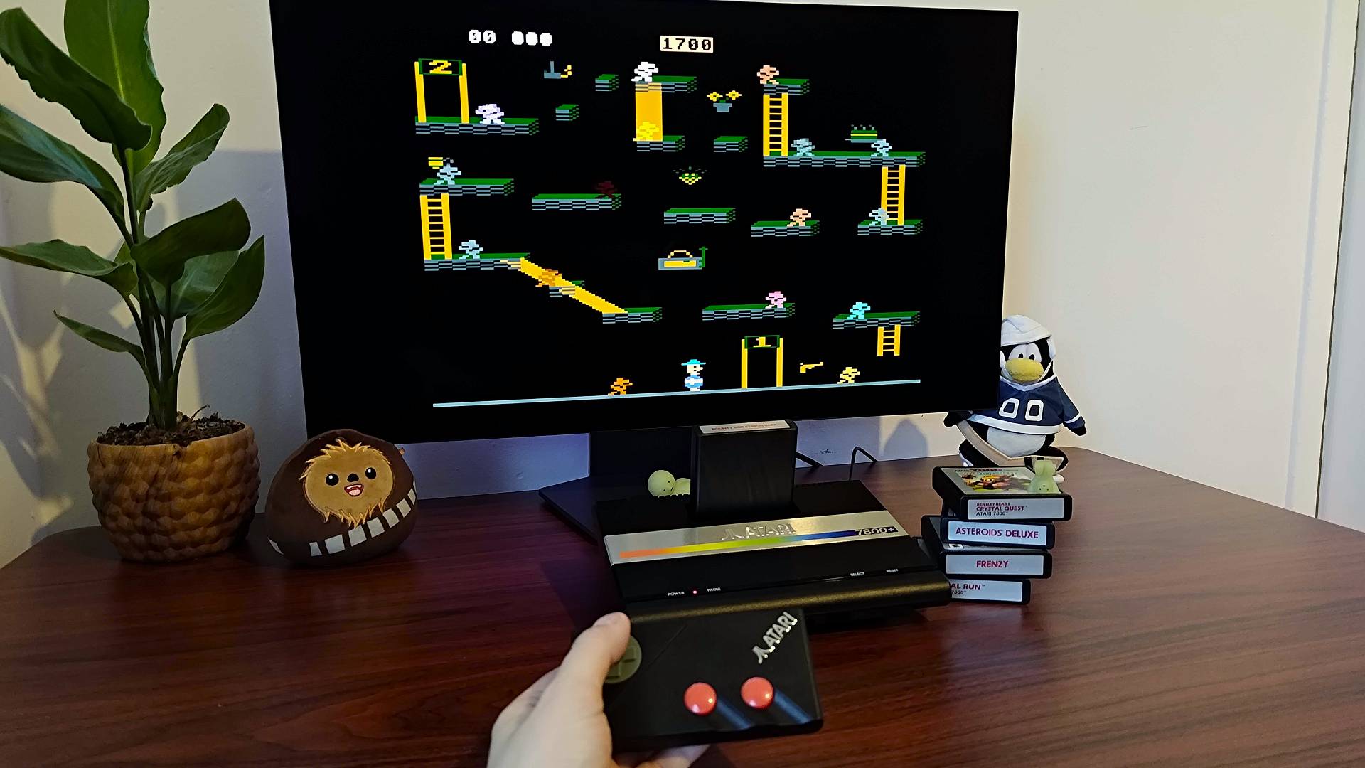 Atari 7800+ connected to monitor with Bounty Bob Strikes Back on screen and hand holding controller infront