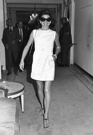 Jackie Kennedy wears a white short dress and thong sandals.