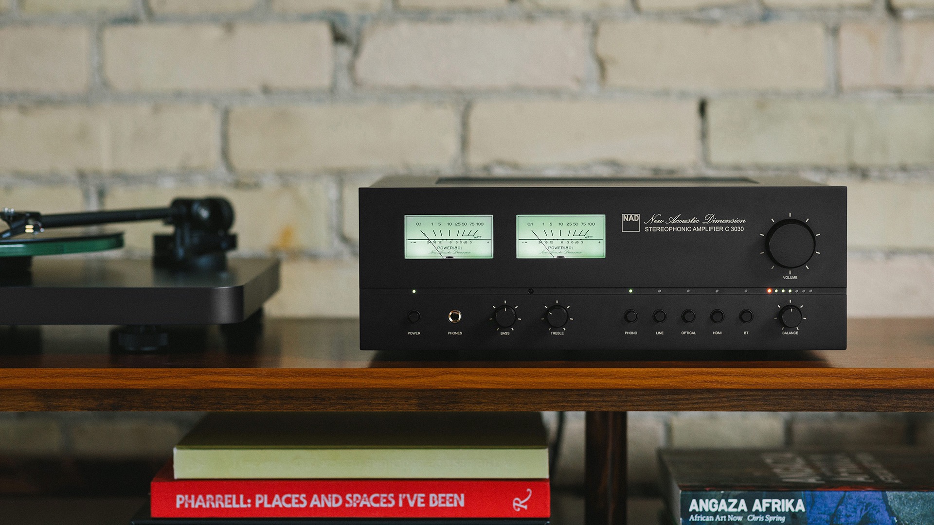 The NAD C 3030 integrated amplifier shot straight-on on a wooden surface next to a record player with books stacked on the shelf below.