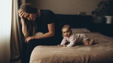 Mother sitting on the bed with her young baby, looking troubled and stressed