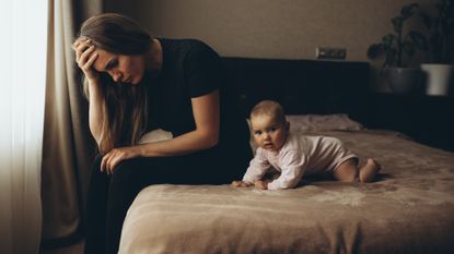 Mother sitting on the bed with her young baby, looking troubled and stressed