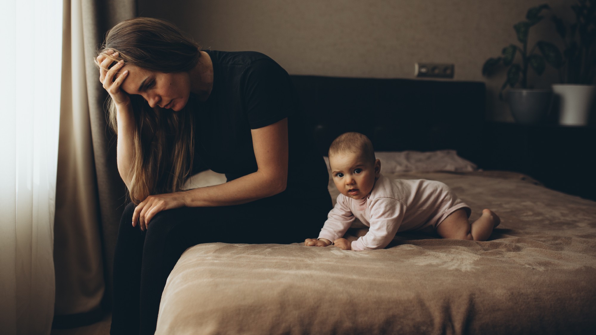 Mother sitting on the bed with her young baby, looking troubled and stressed