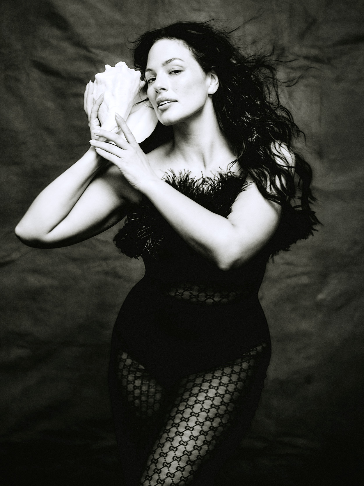 Ashley Graham poses holding a conch shell up to her ear wearing a feather leotard and printed tights in a black and white photo