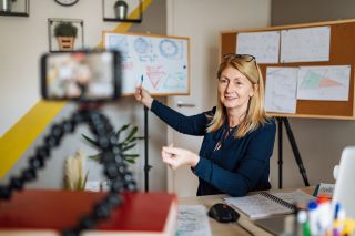 Female teacher points to whiteboard while looking into webcam. 