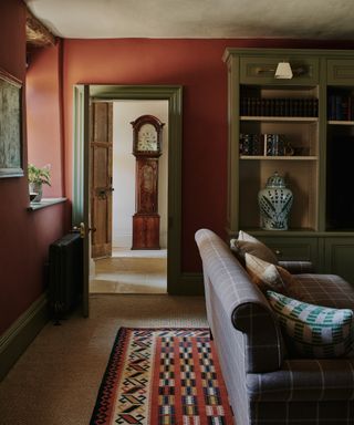 farmhouse style living room with burgundy red walls, sage green woodwork and cabinet, jute carpet, checked couch, rug, Grandfather clock in hallway