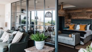 A stylish open-plan bedroom divided by a black metal-framed glass partition. On one side sits a cosy living area with dark armchairs and soft cushions. On the other, a bed with mustard and striped pillows rests against a rustic wood-panelled wall, beneath woven pendant lights. 