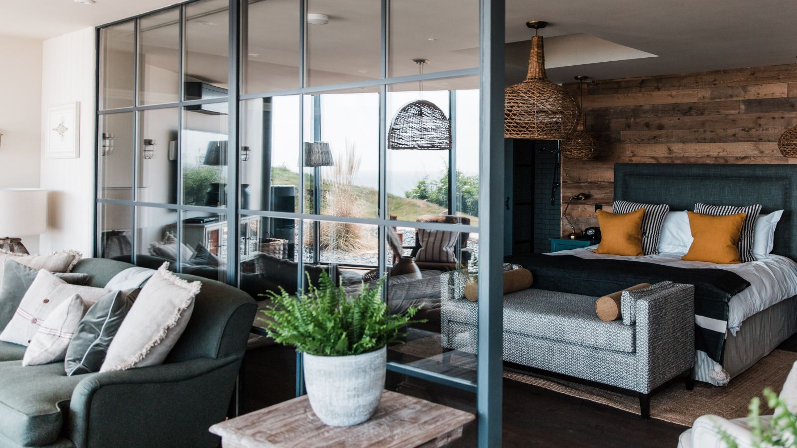 A stylish open-plan bedroom divided by a black metal-framed glass partition. On one side sits a cosy living area with dark armchairs and soft cushions. On the other, a bed with mustard and striped pillows rests against a rustic wood-panelled wall, beneath woven pendant lights. 