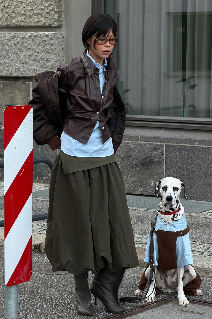 Berlin fashion trends are shown in a street style photo of a woman standing outside beside her Dalmatian dog wearing a brown bomber jacket layered on top of a brown leather button-down shirt and a blue button-down styled with a green ballon skirt, olive green mid-calf ankle boots, and brown sunglasses