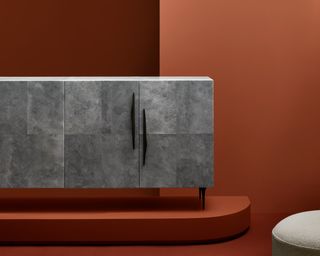 View of a grey cabinet with black handles and legs from the Uncommon Threads collection. The cabinet is on a low plinth in a room with a dark terracotta floor and walls