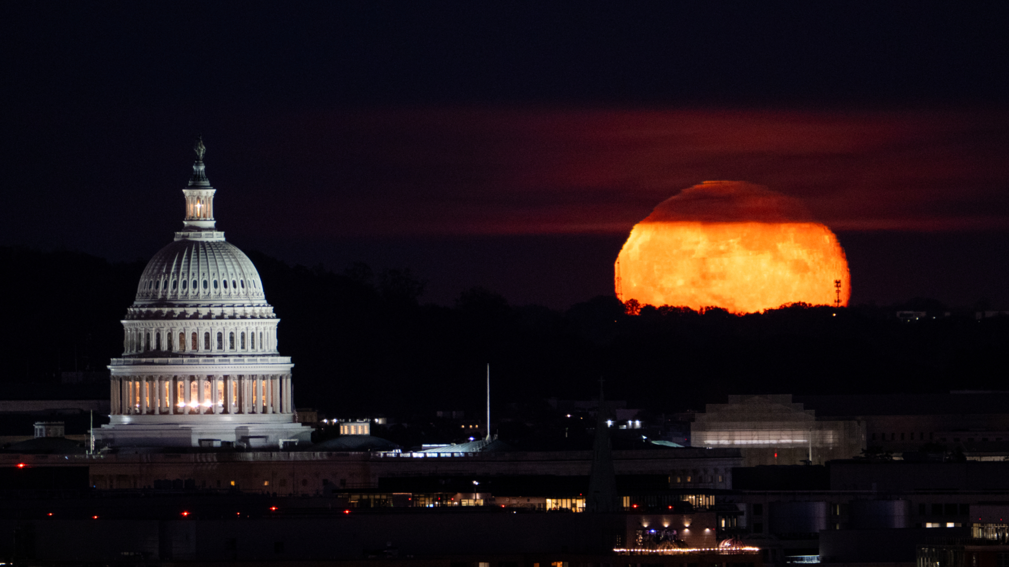 An orange full moon rises with the Capitol Building to its left in Washington D.C. at night.