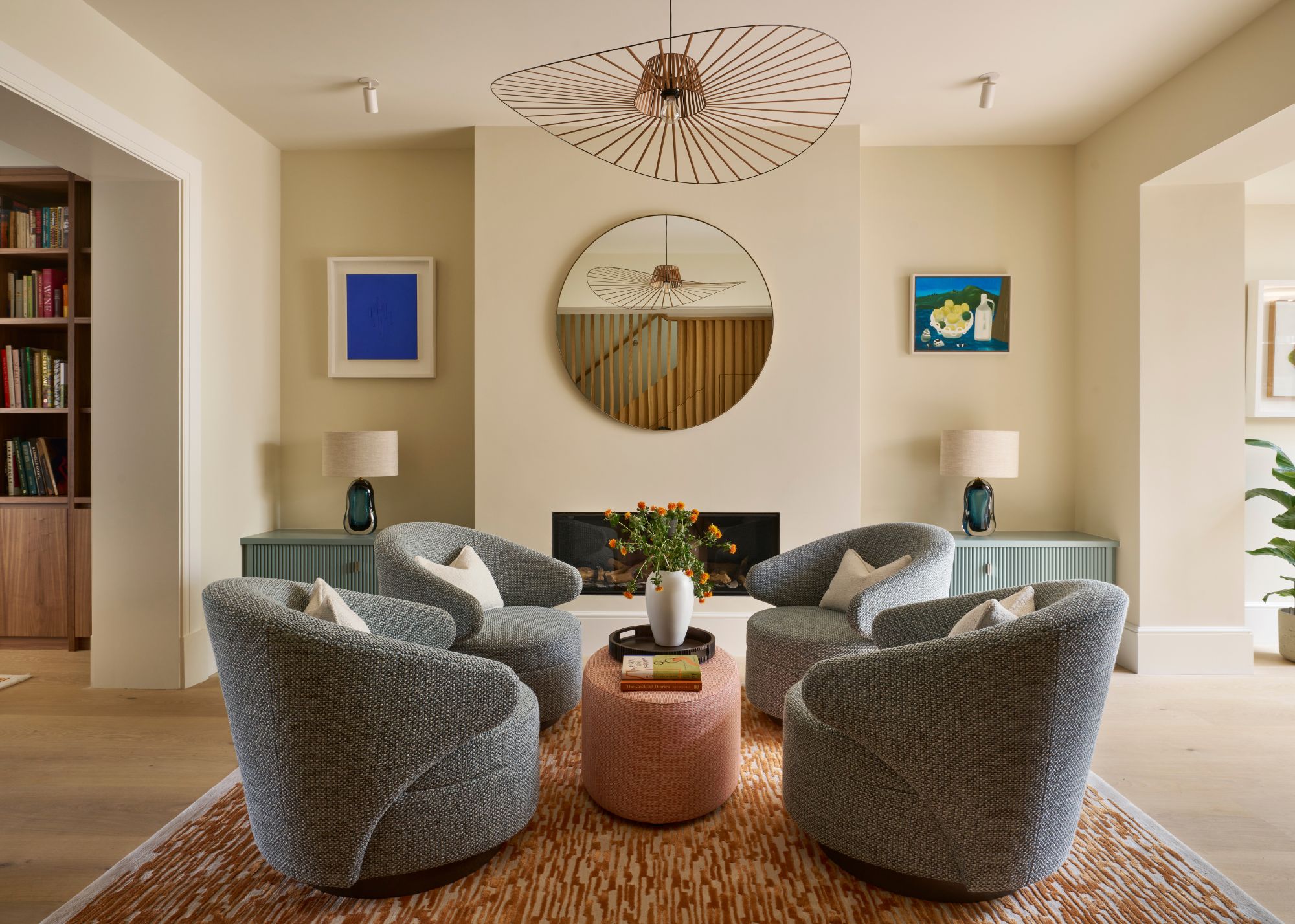 A modern living room with cream walls, a set of four gray armchairs in the middle of the room with a terracotta upholstered coffee table, an orange flecked rug, and a modern pendant light.