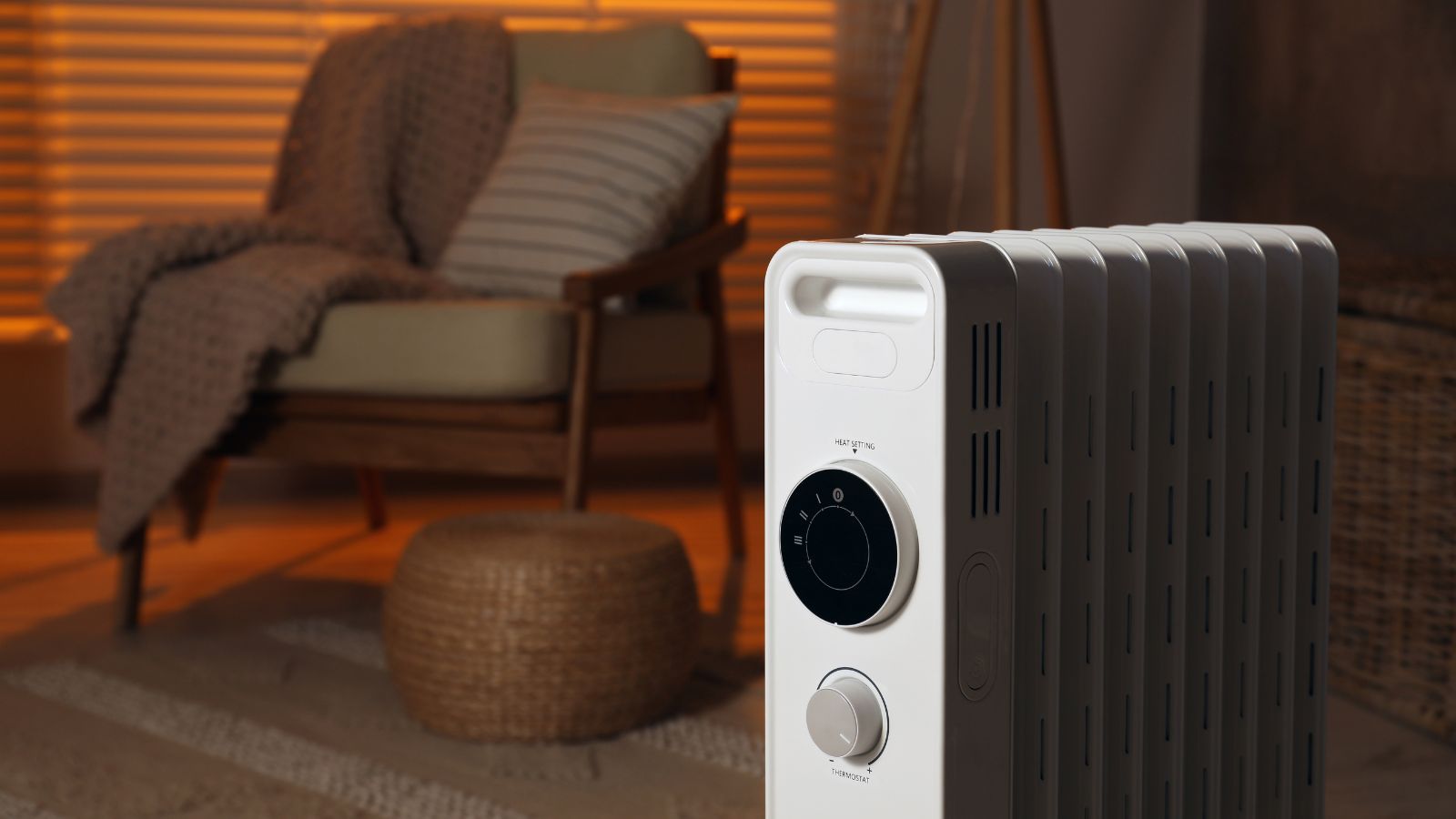 A compact oil-filled radiator stands in the foreground of a softly lit room, its white fins illuminated by warm ambient light.