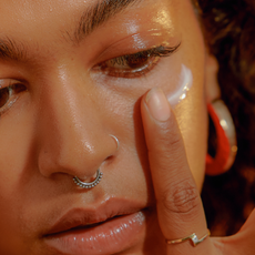 Close up of woman with glowing skin and a nose ring applying skincare to her cheekbone with her index finger