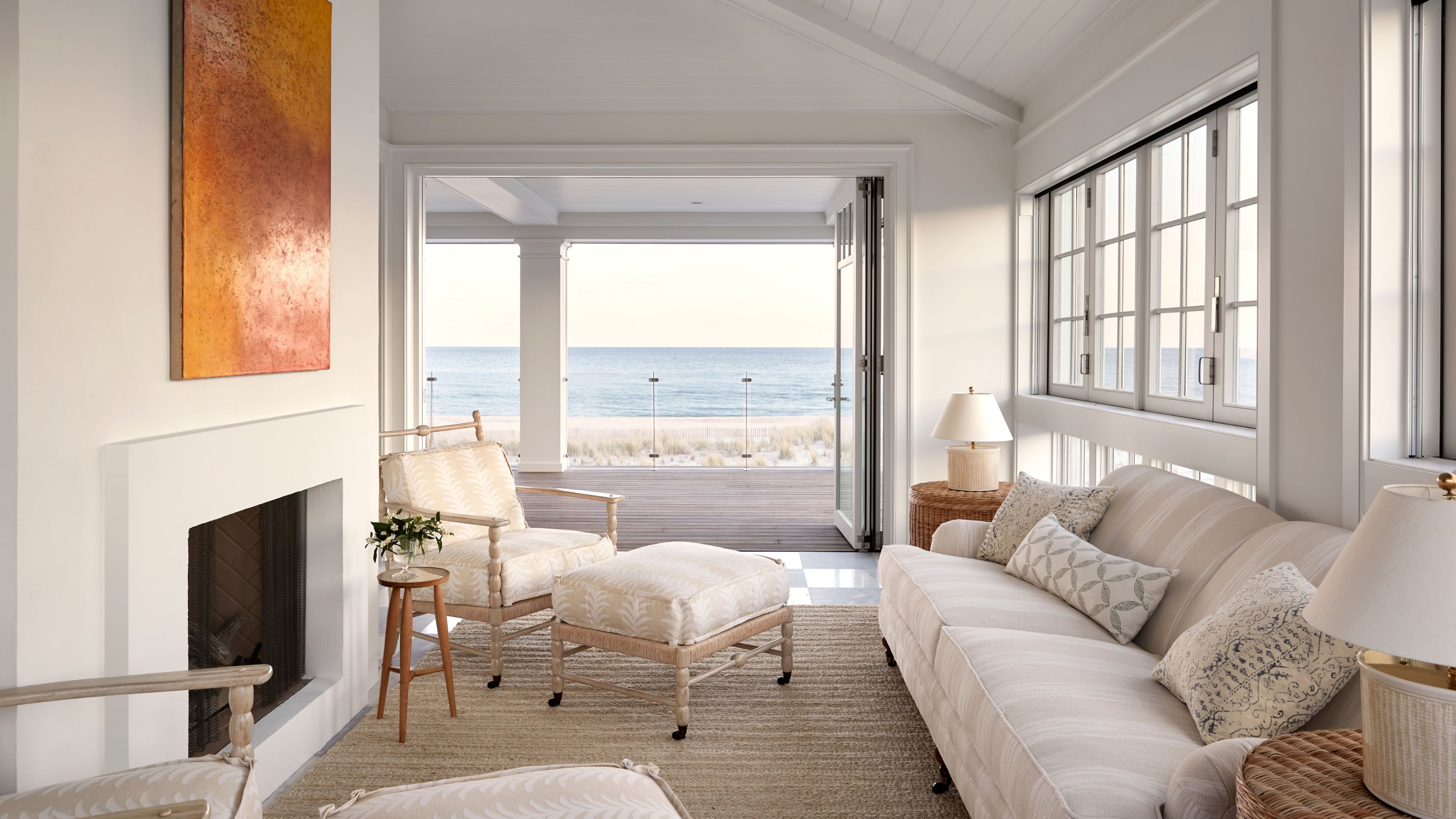 A sun-drenched coastal living room with a white fireplace and a neutral-toned sofa. Large bi-fold glass doors open onto a wooden deck with an expansive view of the ocean and beach at sunset.