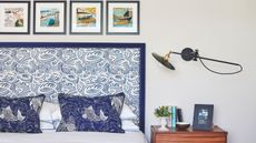 A white bedroom with double bed to the left with statement blue and white patterned headboard, and blue cushions patterned with koi fish. To the right of the headboard is a brown wood bedside table with a small plant, books, a small bowl, and a framed photograph. Above is a wall-mounted angle poise lamp, and above the bed are four small, square framed artworks.